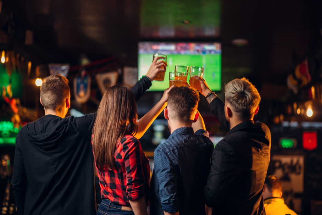 Group of friends standing at a bar watching a sports game on TV.