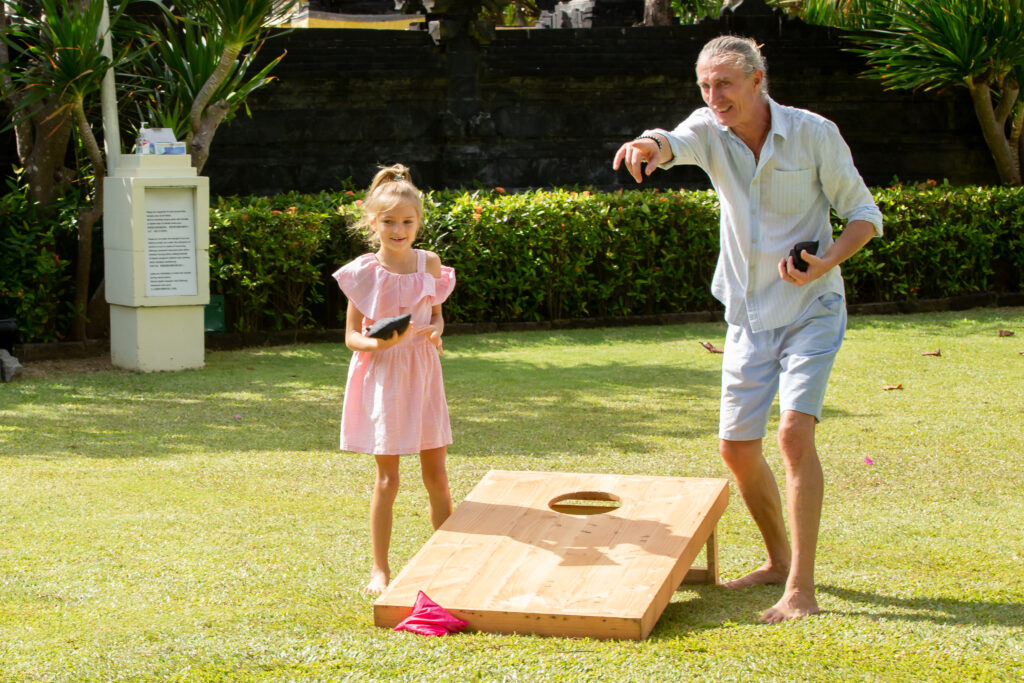 A young girl and an adult are playing cornhole on a sunny lawn.